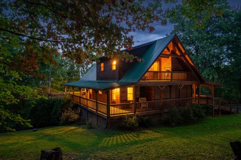 A-frame log cabin with a wraparound porch lit up at twilight, surrounded by autumn trees