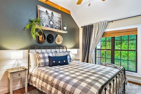 Rustic bedroom with plaid bedding, an iron bed frame, and a large wooded-view window