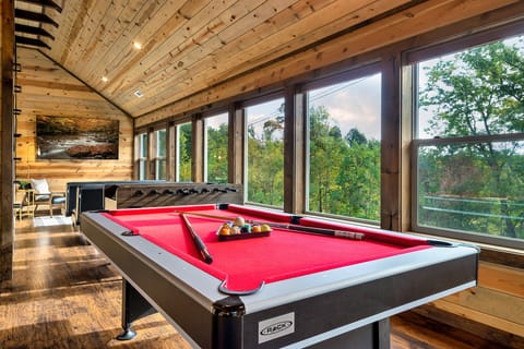 Game room with a pool table and foosball table beneath wall-to-wall windows overlooking treetops