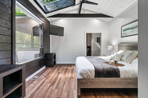 Vaulted-ceiling bedroom with a king bed, wall-mounted TV, and large windows opening to a wooded deck