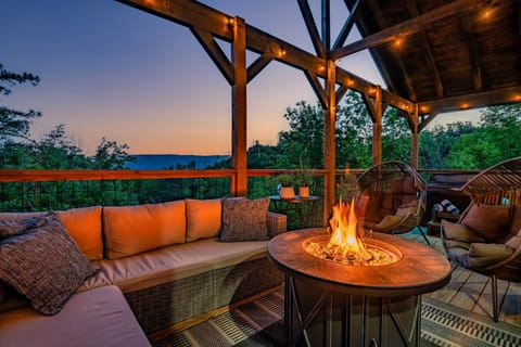 Covered deck lounge with a lit fire pit table and string lights at dusk, mountain view