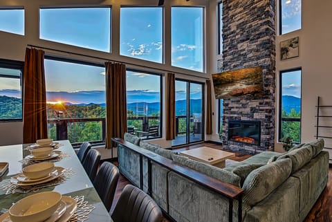 Living and dining area with floor-to-ceiling windows overlooking a mountain sunrise and a stone fireplace