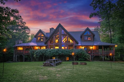Large A-frame log lodge with a wide covered porch and a picnic table on the lawn under a vivid sunset sky