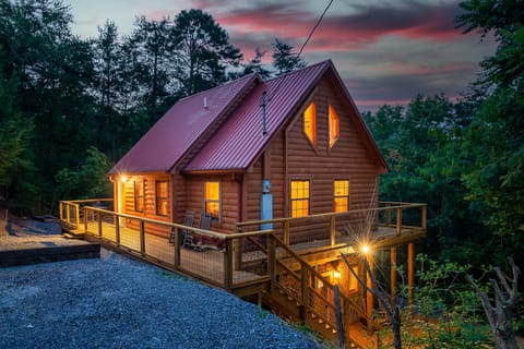 Red-roofed log cabin on a wooded hillside, lit up at twilight with a wraparound deck and rocking chairs