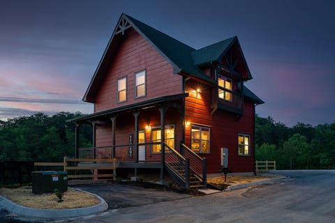 Red two-story cabin with a covered front porch and rocking chairs, lit up at twilight