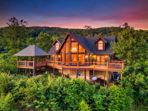 Aerial view of a large A-frame log cabin with a wide covered deck, lit at twilight against a sunset over wooded hills
