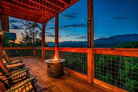 Covered deck with rocking chairs and a lit fire pit table overlooking mountain ridges at twilight
