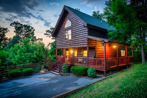 Log cabin with a wraparound porch lit up at twilight, surrounded by trees