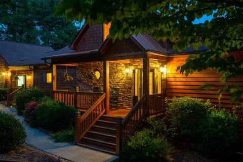 Stone-and-wood cabin entrance with a lit front porch, rocking chairs, and hanging flower baskets at dusk