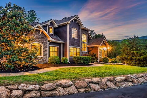 Stone-and-shingle mountain home lit up at twilight with a landscaped lawn and stone garden wall