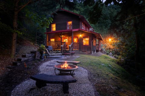 Wooded cabin lit up at dusk with a lit fire pit and stone benches in the foreground