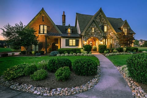 Large stone-and-brick estate home lit up at twilight with manicured landscaping and a curved walkway