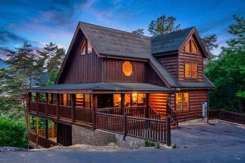 Two-story log cabin with a wraparound covered porch and lit windows at dusk